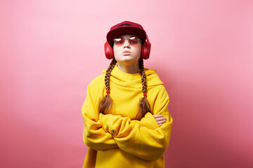 A young girl in a yellow hoodie listens to music with big red headphones. Studio portrait on a pink background.
