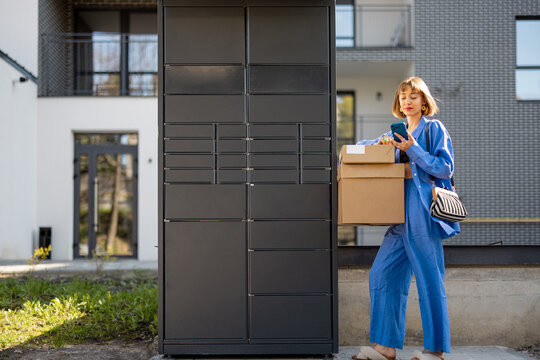 Young woman picks up parcels from automatic post office machine, standing with phone near apartment building. Concept of fast delivery to automatic self lockers
