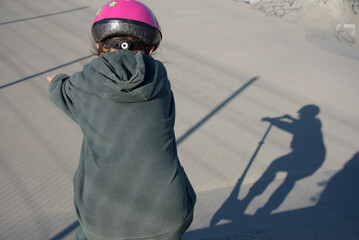 dziecko w różowym kasku przygotowujące się do skoku na hulajnodze, rzucające swój cień w skate parku