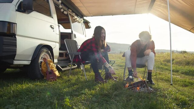 Young caucasian couple roasting marshmallows on the campfire. The romantic road trip, outdoor weekend. Long-haired girl and stylish young man sitting on chairs near a white camper van