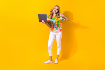 Full length photo of cute confident woman dressed flower print t-shirt showing thumb up working gadget isolated yellow color background