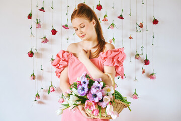 Obraz na płótnie Canvas Studio portrait of pretty young teenage 15 - 16 year old red-haired girl wearing pink coral dress, posing on white background with hanging roses, holding basket with many spring flowers