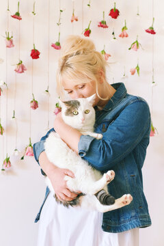 Studio portrait of pretty young teenage 15 - 16 year old girl wearing denim jacket, posing on white background with hanging flowers, holding cute cat, beauty and fashion concept