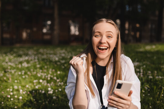 Portrait photo smiling young girl sitting on ground browsing internet with cellphone wearing casual clothes gesturing like winner. Blonde woman look happy, celebrating win, triumph, success.