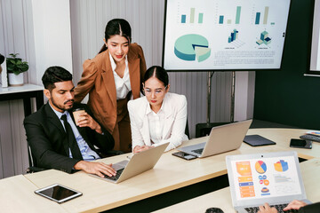 A cheerful and confident Asian businesswoman stands in the back of her office colleagues, presenting bar charts data from a monitor and projector screen. Asian businesswoman leader role at the meeting