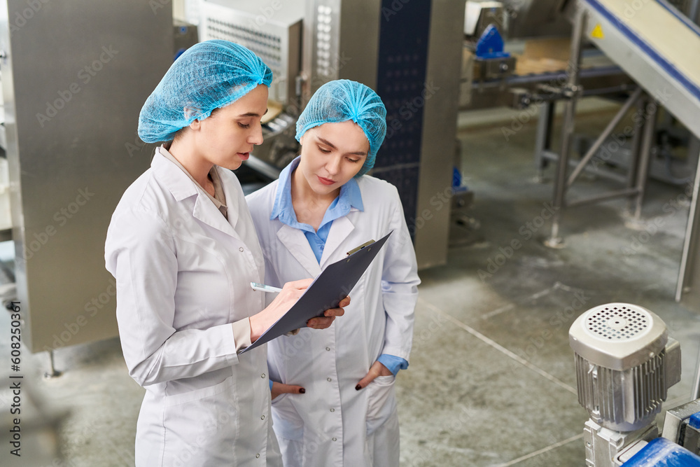 Wall mural concentrated busy food manufacturing workers in caps and lab coats analyzing data in clipboard and f