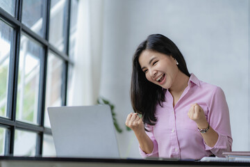 Asian businesswoman rejoices at success in business and raises hands in front of laptop computer Colleagues rejoice at business victories.