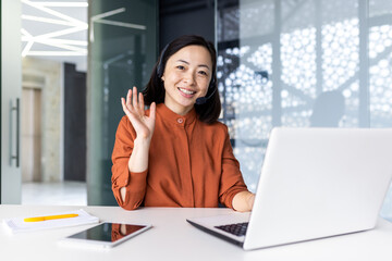 Portrait of successful tech support worker, Asian woman looking at camera, businesswoman with headset for video call, greeting gesture, inside office with laptop.