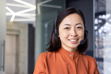 Close up portrait of Asian woman inside modern office with headset for video call, woman smiling...