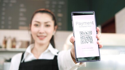 Young Asian woman barista in apron showing QR code for payment on smartphone screen at cafe restaurant. Online payment process. Internet banking. Online wallet. Cashless