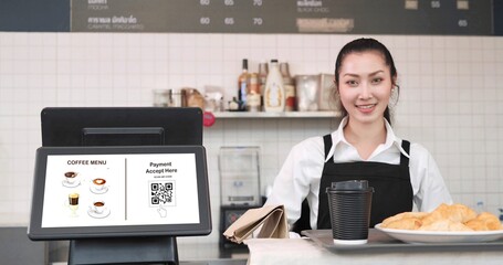 Young Asian woman waitress standing in back counter whlie putting tray with coffee cup and bread, croissants on counter in coffee shop. Menu and QR code payment in screen monitor on counter