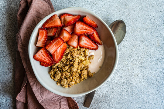 Top view of bowl with granola, yogurt and ripe organic strawberries on grey surface with spoon and napkin
