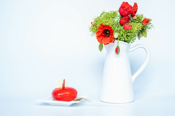 Red heart shaped cake near a white ceramic vase with blooming poppies, on a white background