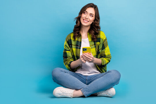 Full length photo of positive person sit floor hold telephone look empty space isolated on blue color background