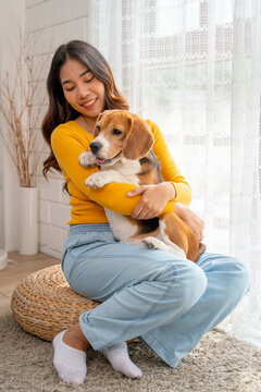 Vertical image of young Asian girl hold and hug beagle dog and sit in front of glass door in her house and she look happy to play fun together.