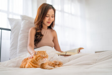 Obraz na płótnie Canvas Side view of pretty Asian woman touch and pat orange cat that lie on bed during she read the book to relax in bedroom with day light.