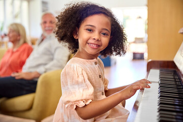 Portrait Of Granddaughter Playing Piano With Loving Grandparents On Sofa At Home In Background