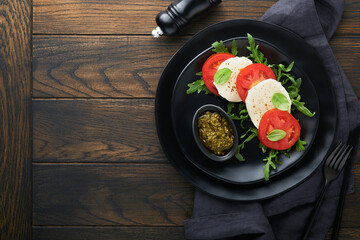 Caprese salad. Italian caprese salad with sliced tomatoes, mozzarella cheese, arugula, basil, olive oil in black plate over old wooden dark background. Delicious Italian food. Top view.