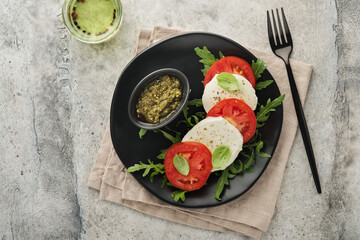 Caprese salad. Italian caprese salad with sliced tomatoes, mozzarella cheese, arugula, basil, olive oil in black plate over old concrete gray background. Delicious Italian food. Top view.