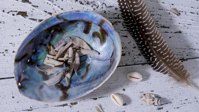 Smudging white sage leaves (Salvia apiana) in an abalone shell with incense feather on old white wood. Overhead view.
