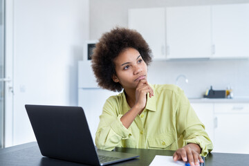 Serious young African business woman, sitting at office workplace with laptop, looking away with...