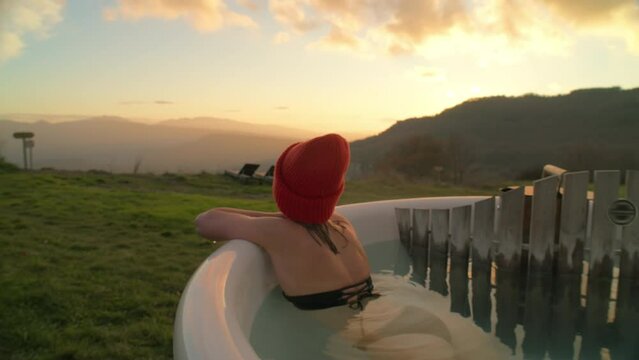 Relaxed and chill young woman look at camera, lay down in steaming hot tub heated by wood burning stove. Cinematic glamping vacation. Retreat in natural spa hotel. 