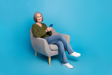 Full length photo of satisfied pleasant woman dressed khaki sweater hold smartphone sit on armchair isolated on blue color background