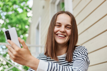 Laughing young woman holding her smartphone in hands while sitting on porch of house using device to send message or browsing internet looking at camera expressing positive emotions.