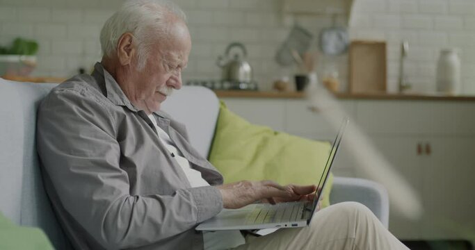 Cheerful Senior Man Using Laptop Typing Working Online Sitting On Sofa At Home Alone. Modern Technology And Elderly People Concept.