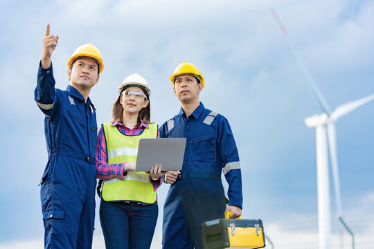 A team of three Asian female mechanics and engineers wearing maintenance suits and vests. holding a tablet to work with toolbox wear a helmet In the background there is a power wind turbine.