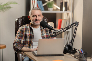 Senior man in checkered shirt is doing podcast. Cheerful blogger with laptop recording video with...