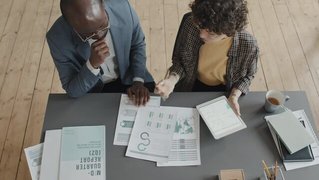 Overhead shot of male African American and female Caucasian coworkers sitting at desk in office at daytime comparing figures in documents and tablet and discussing results