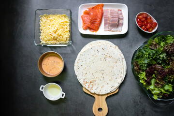 Tortillas with ingredients (smoke salmon, bacon, cheese, tomato, olive oil and sauce on black table background. Homemade delicious tortillas pizza, top view.