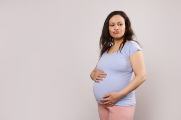 Perplexed pregnant woman expecting baby, holding hands on her big belly in second pregnancy trimester, thoughtfully looking aside, isolated white background. Maternity, childbirth, motherhood concept