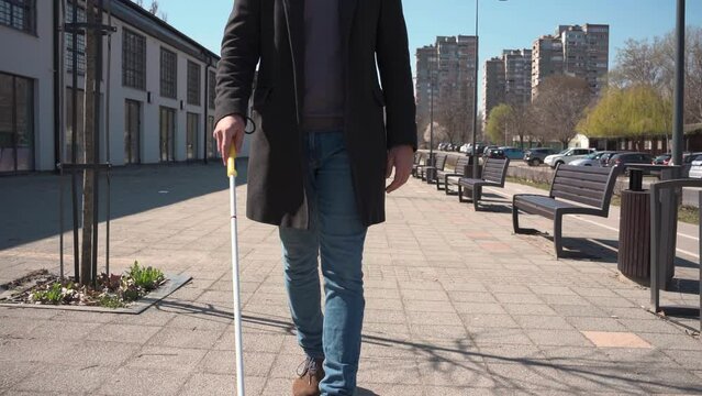 A blind male person using a white cane and walks on city street. 