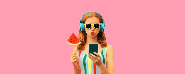 Summer colorful portrait of stylish young woman in headphones listening to music on smartphone with juicy lollipop or ice cream shaped slice of watermelon on pink background