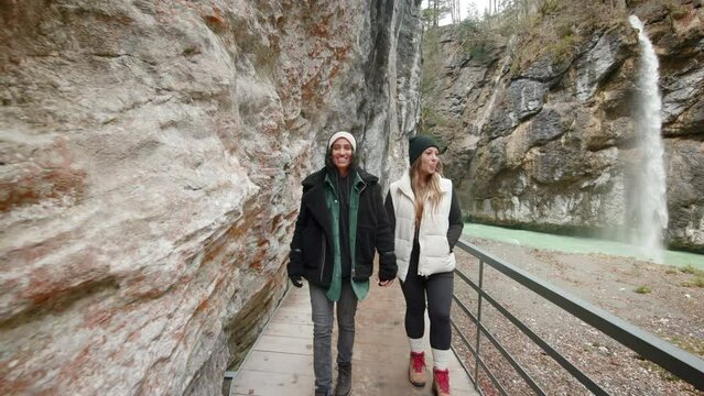 Beautiful walk towards the camera of two LGBT girls, holding hands, amidst the majestic mountains on the wooden bridge adjacent to the enchanting waterfall in Aare Gorge, Switzerland.