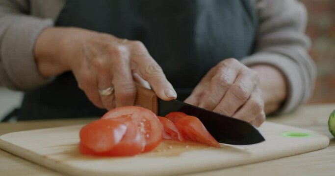 Close-up of senior woman's hands with knife chopping tomato preparing vegetable dish at home. Healthy nutrition and vegetarian meal concept.