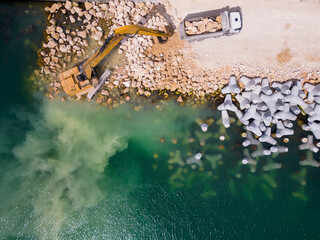 An excavator diligently constructs a dock or breakwater in the sea, its mighty arm reaching out from the shore, creating a resilient structure amidst the waves. Aerial view