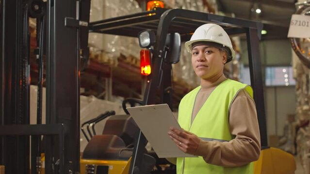 Medium portrait of young Biracial female warehouse worker in white hard hat standing with clipboard against forklift truck with beaming red light