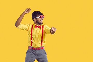 Happy man dancing and having fun. Portrait of funny young Indian guy wearing yellow shirt, red suspenders, bow tie and sunglasses dancing gangnam style isolated on color background. Party concept - obrazy, fototapety, plakaty