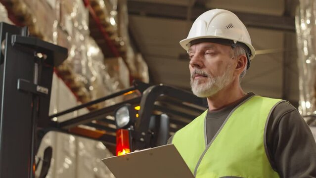 Waist up of Caucasian adult foreman in hard hat looking around big warehouse and taking notes on clipboard while standing against forklift with beaming red light on top