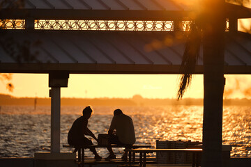 Dark silhouette of people resting under alcove roof on sea shore in public park at sunset