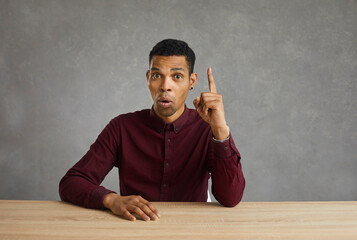 Portrait of a smart confident dark-skinned young man who has a great innovative idea pointing his finger at the sign of eureka. Guy shows index finger up sitting at the table on a gray wall background