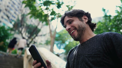 Fototapeta premium One joyful young handsome man conversing while holding smartphone device and standing outside in city park