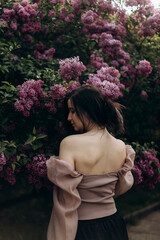 young beautiful brunette woman stands with her back near a blooming lilac. a girl in a beige blouse with an open back stands in a lilac garden