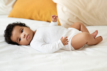 Serious black little child girl or boy in clothes lies on white comfort bed in bedroom. Health care, newborn