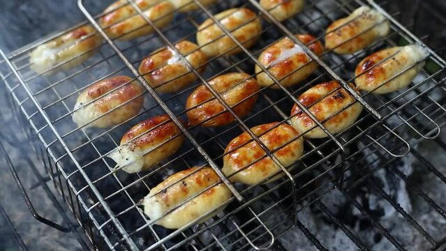 Small grilled sausages on grill with smoke and flame on dark charcoal background outdoor. Baked ruddy sausage grilled on a grill grate. The concept of a picnic in nature. Video fottage in HD 25 FPS