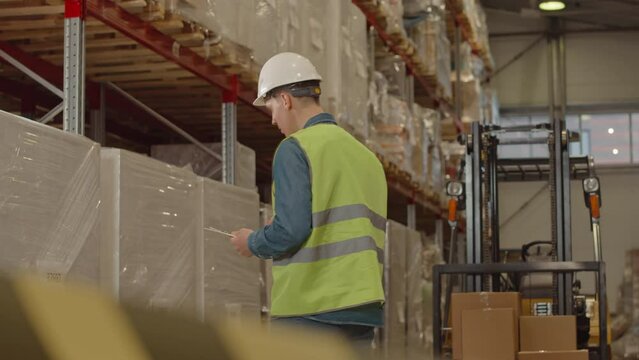 Medium shot of young Caucasian male warehouse worker wearing hard hat examining cardboard boxes on pallets and taking notes on clipboard while doing inventory management during shift