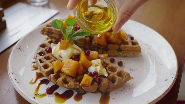 Woman pouring honey over fresh vegetarian waffles with pumpkin in a cafe, close-up hand